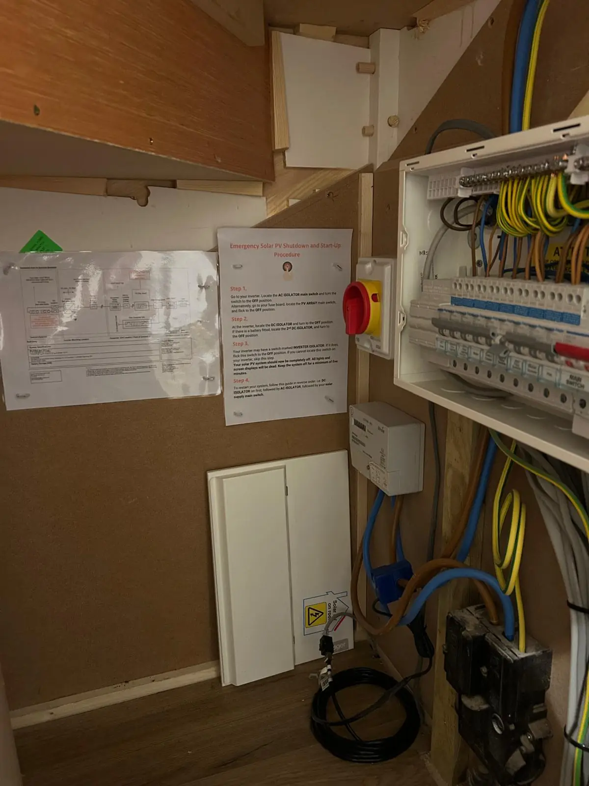 A consumer unit in a cupboard with the cover off, showing tidy grouped wiring, alongside a wall mounted PV emergency shutdown notice.