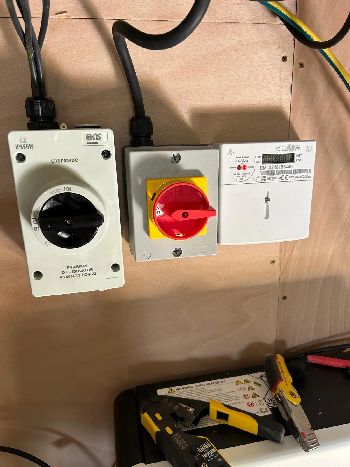 A PV array DC isolator, a rotary AC isolator and a generation meter mounted in a row on a plywood board, with hand tools on the shelf below.