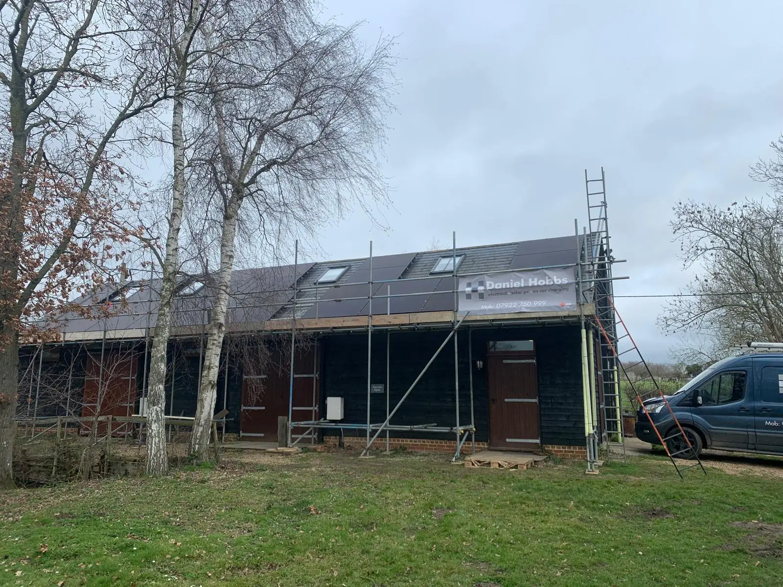 A black timber clad barn conversion under scaffolding, with solar panels being installed across the roof slope and a Daniel Hobbs Electrical banner on the scaffold.