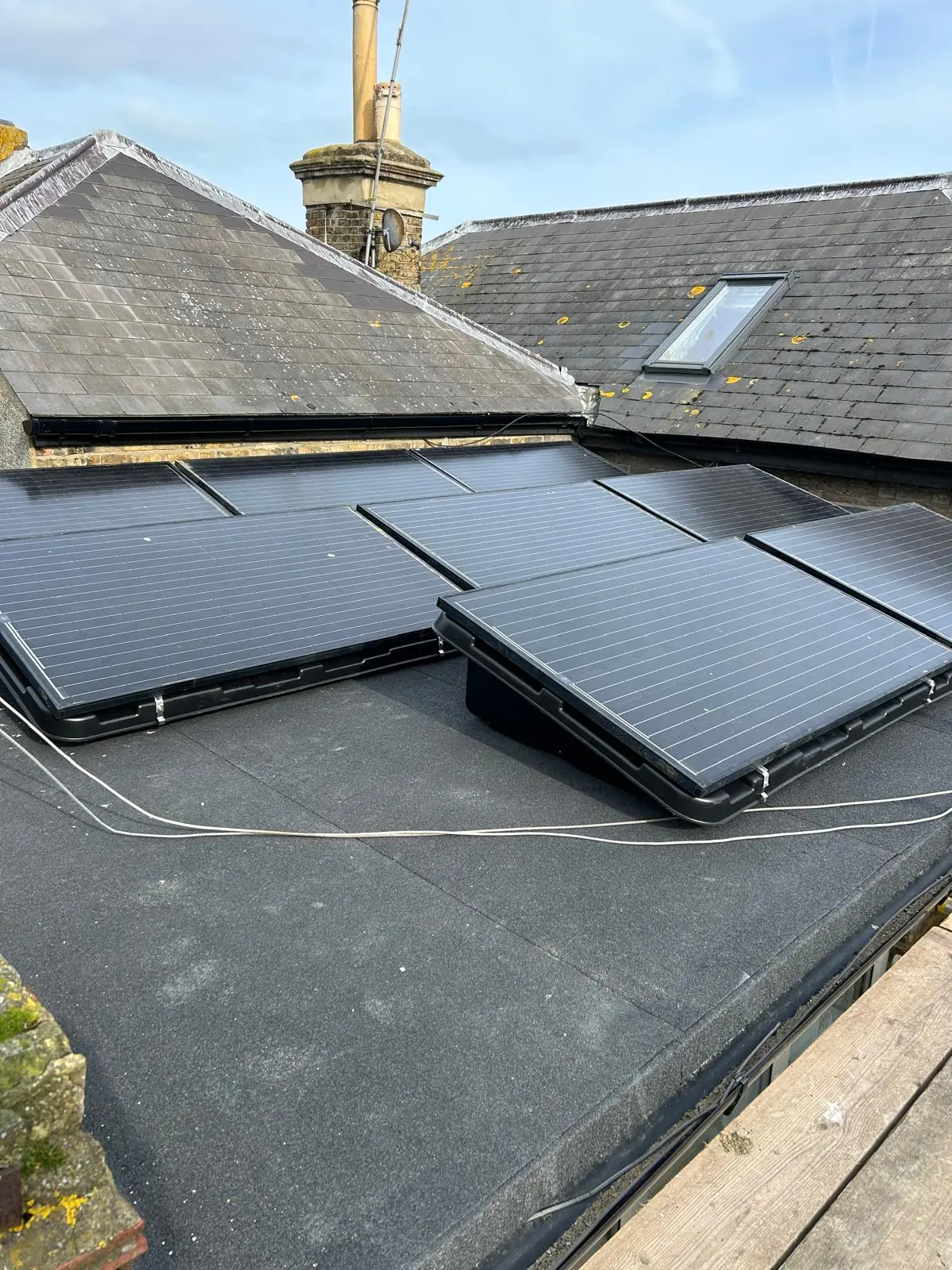 Tilted solar panels on a flat roof beside a stone chimney and slate roof slopes, with DC cabling run across the covering.