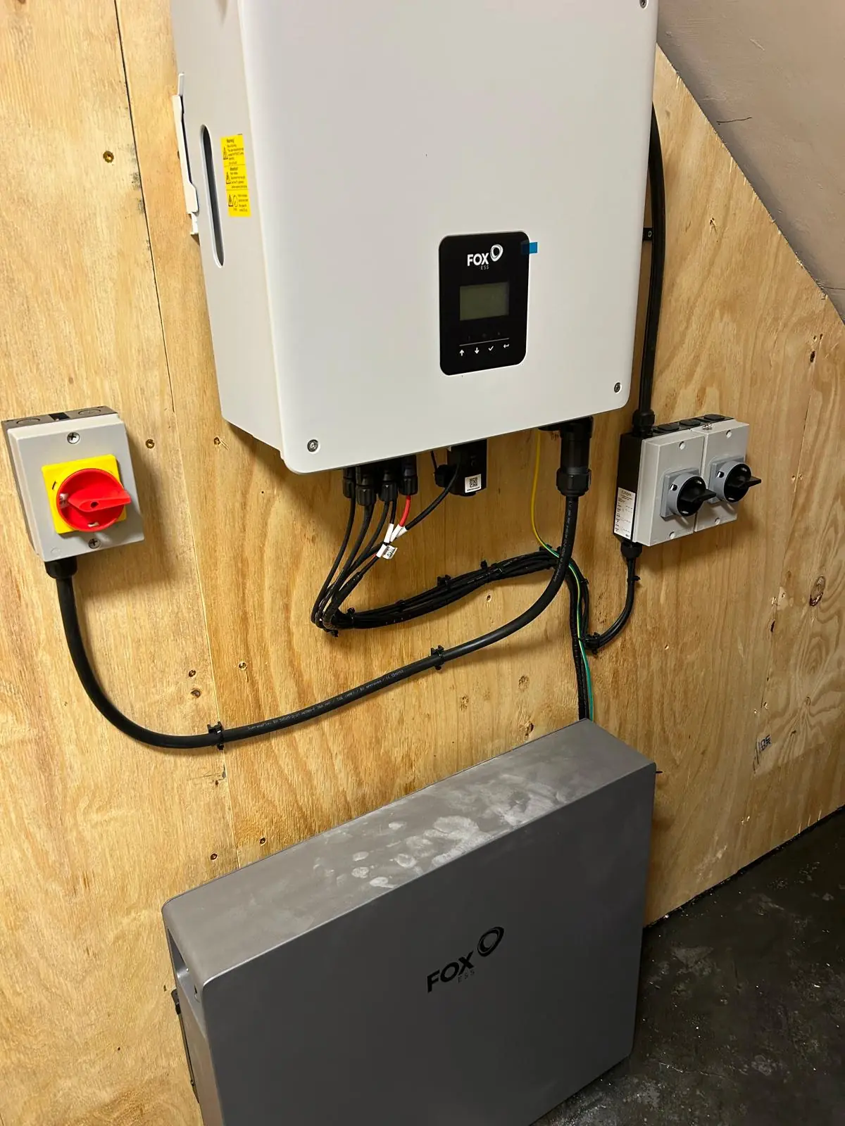 A wall mounted solar inverter and battery unit on a plywood board, with an AC isolator, a DC isolator and cabling run between them.