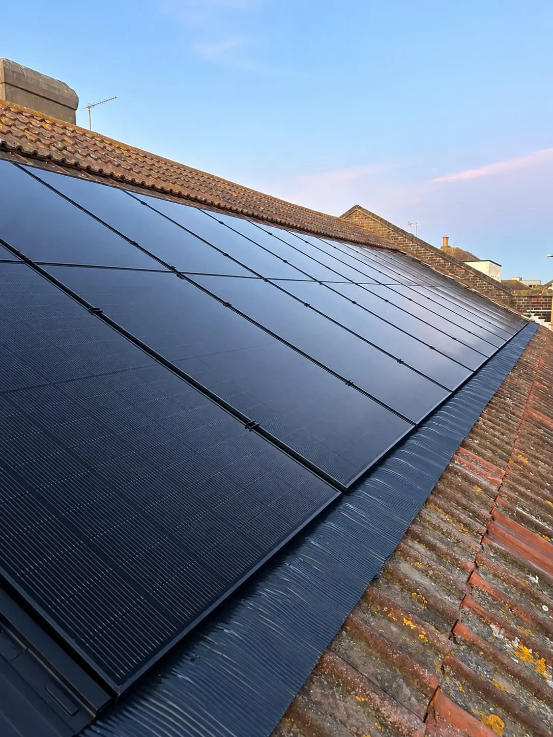 A finished all black array across a pantile roof at dusk, with rooftops and a chimney behind.